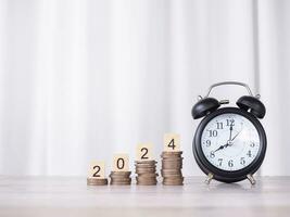 Black alarm and Wooden block with number 2024 on stack of coins. The concept of saving money, Financial, Investment and Business growing in new year 2024. photo