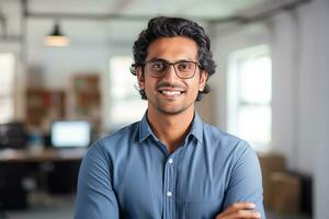 ai generado un joven indio masculino diseñador, ingeniero, arquitecto quien es vistiendo lentes y un azul camisa sonriente en pie en el oficina y mirando a el cámara foto