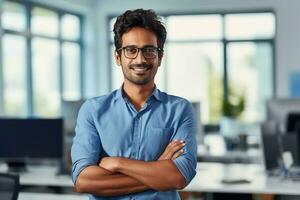 ai generado un joven indio masculino diseñador, ingeniero, arquitecto quien es vistiendo lentes y un azul camisa sonriente en pie en el oficina y mirando a el cámara foto