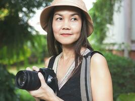 mujer asiática, con sombrero y blusa negra sin mangas, parada en el jardín y sosteniendo una cámara dslr, sonriendo alegremente. foto