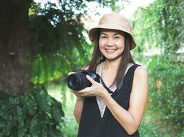 mujer asiática, con sombrero y blusa negra sin mangas, parada en el jardín y sosteniendo una cámara dslr, sonriendo alegremente. foto