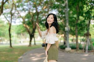 retrato de asiático joven mujer viajero con Costura sombrero y cesta y un cámara en verde público parque naturaleza antecedentes. viaje viaje estilo de vida, mundo viaje explorador o Asia verano turismo concepto. foto