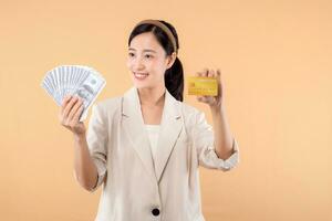 portrait of happy successful confident young asian business woman wearing white jacket holding cash money dollars and credit card standing over beige background. millionaire business, shopping concept photo