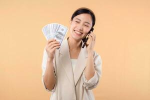 portrait of happy successful confident young asian business woman wearing white jacket holding smartphone and cash money dollar standing over beige background. millionaire business, shopping concept. photo