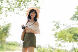 Portrait of asian young woman traveler with weaving hat and basket and a camera on green public park nature background. Journey trip lifestyle, world travel explorer or Asia summer tourism concept. photo