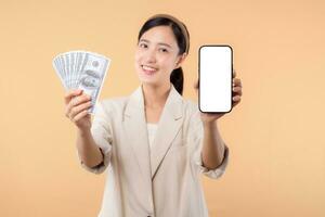 portrait of happy successful confident young asian business woman wearing white jacket holding smartphone and cash money dollar standing over beige background. millionaire business, shopping concept. photo