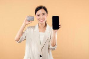 portrait of happy successful confident young asian business woman wearing white jacket holding smartphone and credit card standing over beige background. shopping concept. photo