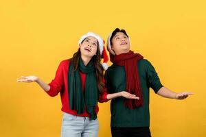 Photo of an excited Asian couple in Christmas clothes looking up with open hands on a yellow studio background.