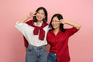 Two happy Asian female friends showing v sign on pink background. and looking at the camera photo