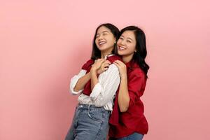 Two happy smiling Asian female friends hugging each other, wearing red and white shirts, standing isolated on pink background. photo