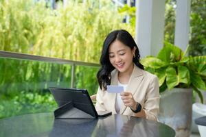 Attractive young Asian freelancer woman with tablet and credit card doing online shopping in the outdoor seating area of a coffee shop. photo