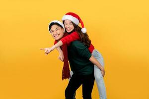 Smiling Asian couple wearing sweaters and Santa hats playfully posing piggyback on a yellow background. Happy New Year or Christmas holiday concept. photo