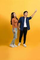 Full view of young Asian couple in casual clothes pointing at copy space with hands isolated on yellow background. photo