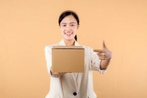 retrato de contento exitoso confidente joven asiático negocio mujer vistiendo blanco chaqueta participación paquete o empaquetar caja aislado en beige estudio fondo, entrega mensajero y Envío Servicio concepto. foto