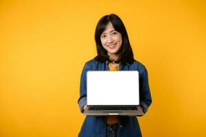 portrait young happy woman wearing yellow t-shirt and denim shirt holding laptop and point finger to screen isolated on yellow studio background. business technology application communication concept. photo