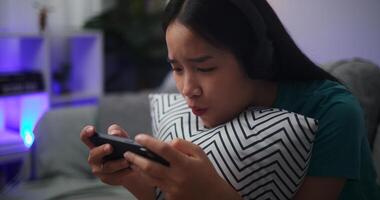 retrato de joven asiático mujer vistiendo lentes y auriculares disfruta jugando en línea deporte juegos en teléfono inteligente sentado en sofá en el vivo habitación a hogar, jugador estilo de vida concepto. foto