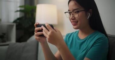 retrato de joven asiático mujer vistiendo lentes y auriculares disfruta jugando en línea juegos en su teléfono inteligente en el sofá en el vivo habitación, jugador estilo de vida concepto. foto