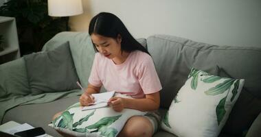 retrato de joven asiático mujer sentado en un sofá participación financiero cuentas y tomando notas en un cuaderno a hogar Hogar Finanzas foto