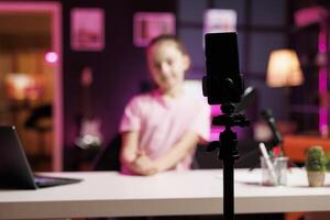 Young child in blurry background doing livestream using smartphone on professional recording tripod. Cute kid does live broadcast using cellphone camera to talk with online audience, focus on phone photo