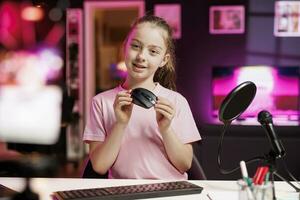 Cute girl in studio reviewing gaming mouse in front of camera in neon lit studio. Joyous youngster filming electronics haul, presenting wireless computer peripheral to viewers photo