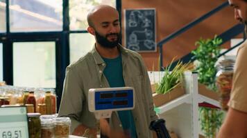 Merchant using scale at checkout, weighting food supplies and fresh produce for middle eastern buyer in grocery store. Vendor selling pantry items in bulk to man in local supermarket. Handheld shot. photo