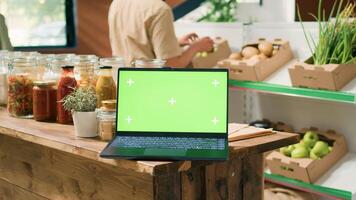 Greenscreen laptop in local eco store with crates of organic homegrown produce and bulk products in jars on shelves. PC running isolated display with mockup copyspace template. photo