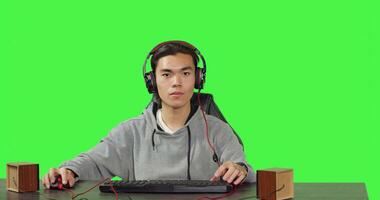 POV of asian man plays pc videogames at desk, sitting against greenscreen background. Gamer having fun with friends online on gaming contest, wearing headphones at workstation. photo