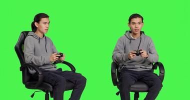 Asian guy playing videogames over greenscreen backdrop, using controller and having fun with friends on online gaming competition. Young man gamer enjoying rpg gameplay, blank background. photo