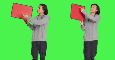 Young man holds speech bubble in studio, presenting blank copyspace on cardboard icon against greenscreen. Asian person working on creating marketing presentation, advertisement. photo