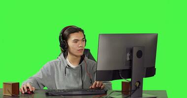 Asian gamer on desktop at workstation, playing internet videogames with others. Young man having fun with role playing battle, communicating with friends over greenscreen template. photo