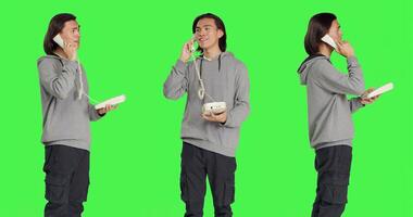 Asian person talking on landline phone, answering call on telephone with cord while he stands against greenscreen backdrop. Young adult using retro phone for remote communication. photo