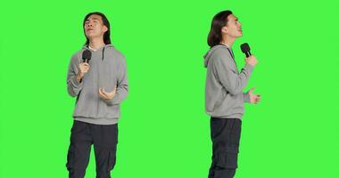 Trendy guy performs karaoke on camera, using recording device to sing popular melodies and lyrical content. Young man having an excellent time over greenscreen backdrop, creating sounds and songs. photo