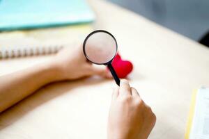 Closeup hands of medical student holding a magnifying glass on a yarn red heart and blurred background. Asian medical student with Heart disease concept. photo