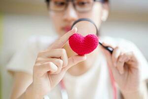 Closeup medical student holding a magnifying glass and looks at a yarn red heart on blurred background. Asian medical student with Heart disease concept. photo