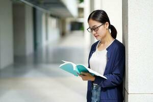 de cerca asiático joven hembra estudiante en casual paño leyendo un libro de texto para el examen a colegio edificio y borroso antecedentes. asiático colegio concepto. foto