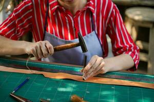 Closeup and crop hands of young female leather maker using a small hummer with leather punching steel to make a leather belt for customers. photo