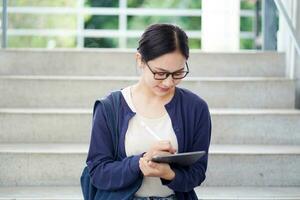 Closeup Asian young female student in casual cloth reading and make a short note on tablet for the exam on the stairs of the school building and blurred background. Asian school concept. photo