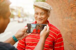 Closeup back view of a man take a photos to senior Chinese tourist by smart phone while walking around the city on blurred of city and sun flare background. Senior tourist concept