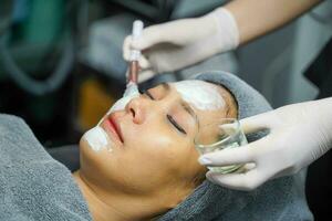 Closeup cosmetologist is applying facial white mask cream on woman client face in beauty clinic. photo