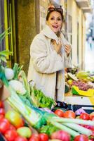 Smiling woman in warm coat standing by stall of fruits and vegetables photo