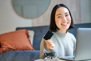 Happy smiling girl with credit card, paying her bills online on computer, doing shopping on her laptop, sitting at home photo