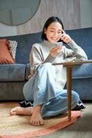Vertical shot of stylish korean woman sitting on floor at home, using her mobile phone, holding smartphone and smiling photo