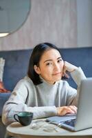 Vertical shot of young asian woman looking tired, smiling with exhausted expression, working from home on her laptop photo