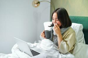 Hardworking asian girl staying in bed with cold, catching flu but working from home with laptop, studying while feeling sick photo