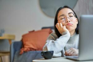 Portrait of korean girl sits bored, student looks gloomy at laptop, sitting at home and expressing boredom photo