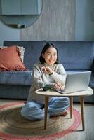 Vertical shot of asian girl sits on floor at home, working on laptop, studying at cozy place, using computer to freelance photo