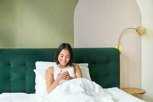 Portrait of smiling asian woman lying in bed, looking at smartphone, using mobile phone with happy face, relaxing on lazy morning photo