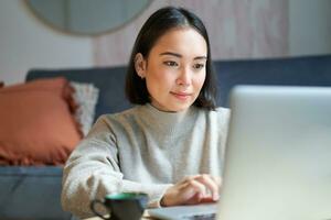 yo empleado joven coreano mujer trabajando en remoto, mecanografía en computadora portátil, estudiando a hogar en vivo habitación foto