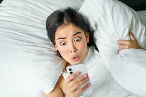 Portrait of asian girl wakes up in morning, looks at mobile phone with shocked face, overslept, hugging pillow and grimacing photo