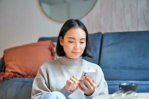 retrato de sonriente asiático mujer se sienta a hogar, usos teléfono inteligente aplicación, pedidos entrega, mensajes alguien en móvil teléfono solicitud foto